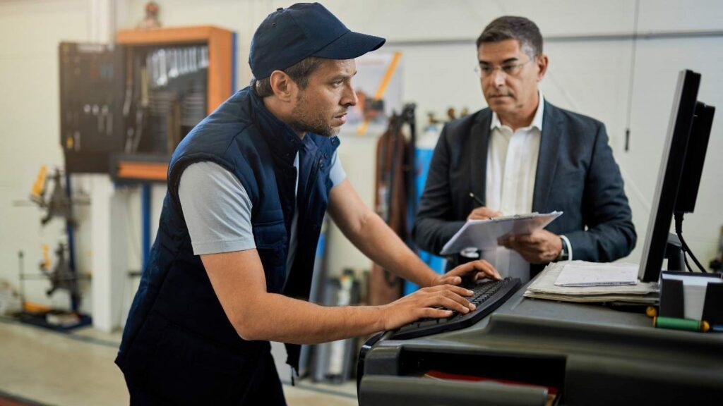 mid adult inspector overseeing auto repairman who is working n computer workshop focus is mechanic (1)